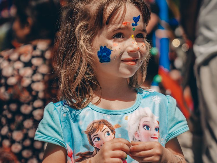 Image of A young girl wearing a blue Frozen dress with Anna and Elsa on it stands outdoors, her face decorated with stickers and colourful paint, smiling softly. People and festive decorations are blurred in the background.