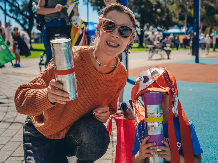 Image of A woman and child kneel on a playground, smiling and holding homemade percussion instruments made from tins. Other people and children are in the sunny park background, enjoying an outdoor event.