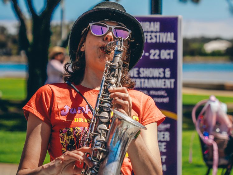 Image of A person wearing a hat, sunglasses, and a red T-shirt plays the saxophone outdoors during a sunny festival, with a purple event sign and park scenery in the background.
