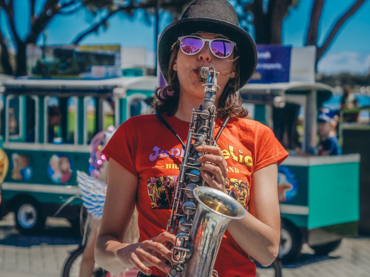 Image of A woman wearing sunglasses and a black hat plays a saxophone outdoors. She has on a red T-shirt, and there is a green tram and people in colourful costumes in the background.
