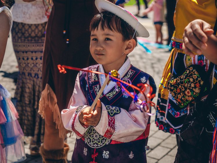 Image of A young boy in traditional festive clothing holds colourful ribbon sticks, surrounded by people in vibrant outfits, celebrating outdoors on a sunny day.