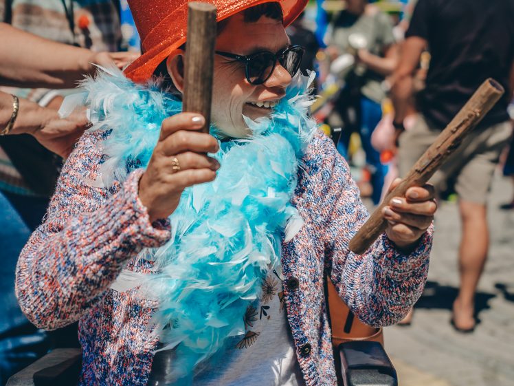 Image of A person in a wheelchair wearing a sparkling orange hat, sunglasses, a blue feather boa, and a colourful jumper smiles whilst holding two sticks at an outdoor event, surrounded by other people.