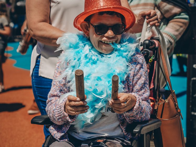 Image of A person in a wheelchair wearing a sparkly orange hat, sunglasses, feather boa, and holding rhythm sticks, smiles at an outdoor event. Another person stands behind them, and a woman in fancy dress is in the background.