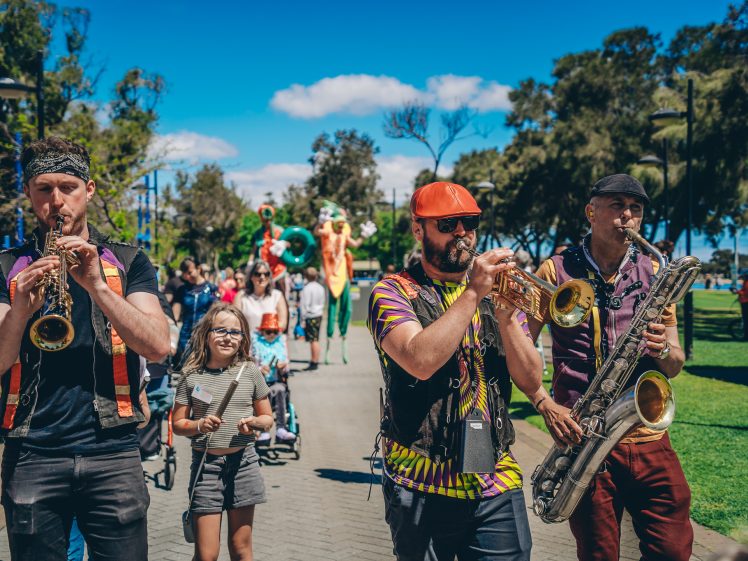 Image of A lively street band plays brass instruments as they parade through a park, joined by a smiling girl with a striped jumper. Other people and performers on stilts are visible in the background under a sunny sky.