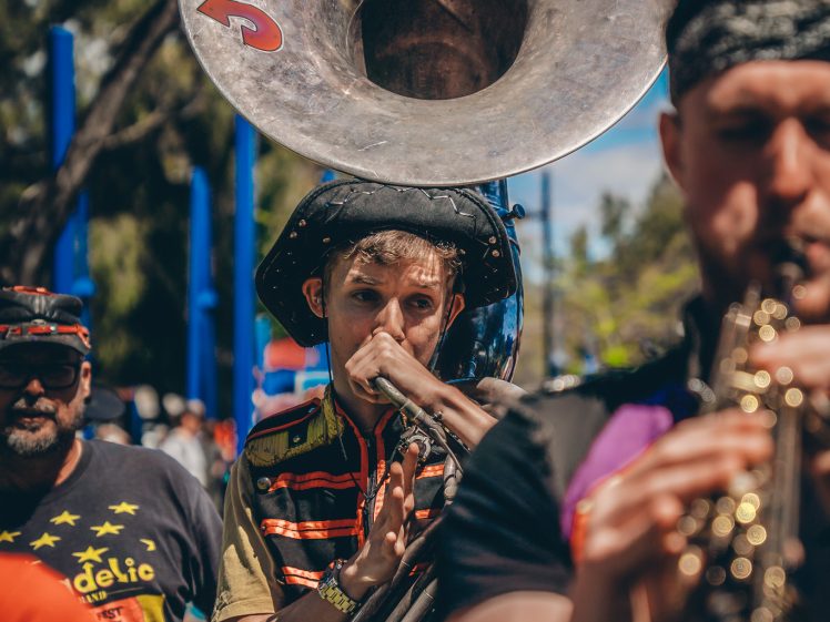 Image of A musician plays a sousaphone labelled Junkadelic in an outdoor setting, surrounded by other band members playing instruments under a bright blue sky.