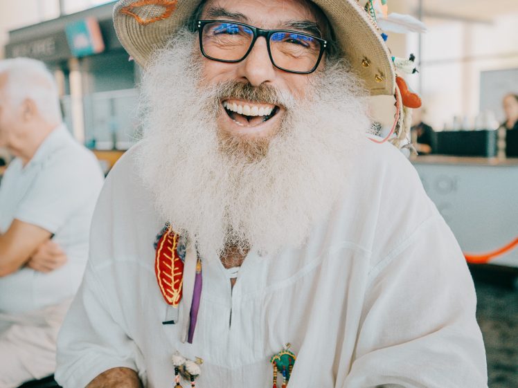 Image of A smiling person with a long white beard and glasses wears a colourful, decorated hat and a white shirt. They have multiple bracelets and badges, sitting indoors with natural light and people in the background.