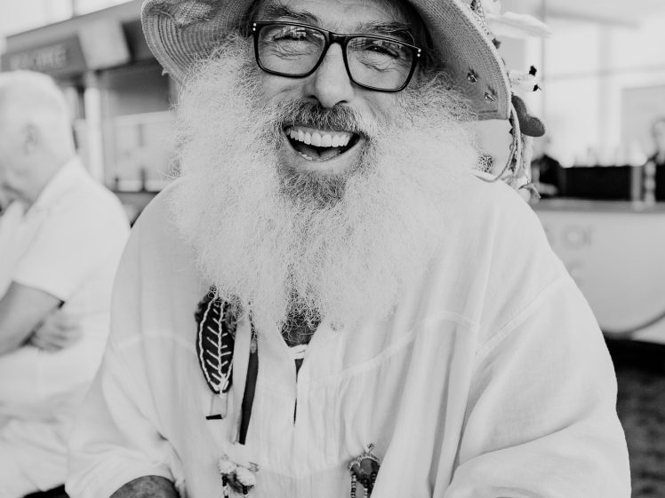 Image of A cheerful older man with a large white beard and glasses smiles whilst wearing a decorated hat with feathers and trinkets, sitting indoors at a table. The image is in black and white.