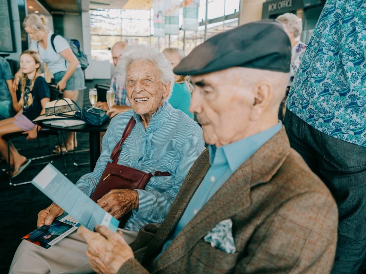 Image of An elderly woman smiles at the camera whilst sitting next to an elderly man reading a leaflet. They are indoors with several people in the background.