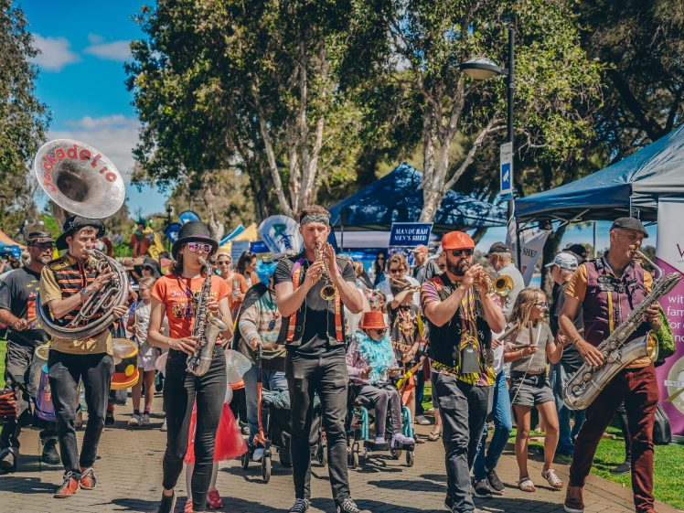 Image of A lively outdoor parade features a colourful brass band playing instruments as they walk through a park, surrounded by people, market stalls, and trees on a sunny day.