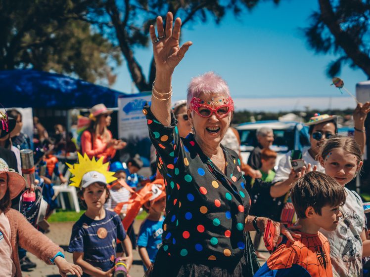 Image of A joyful elderly woman in colourful polka-dot clothing and red glasses waves whilst leading a lively group of children in fancy dress during an outdoor festival on a sunny day.