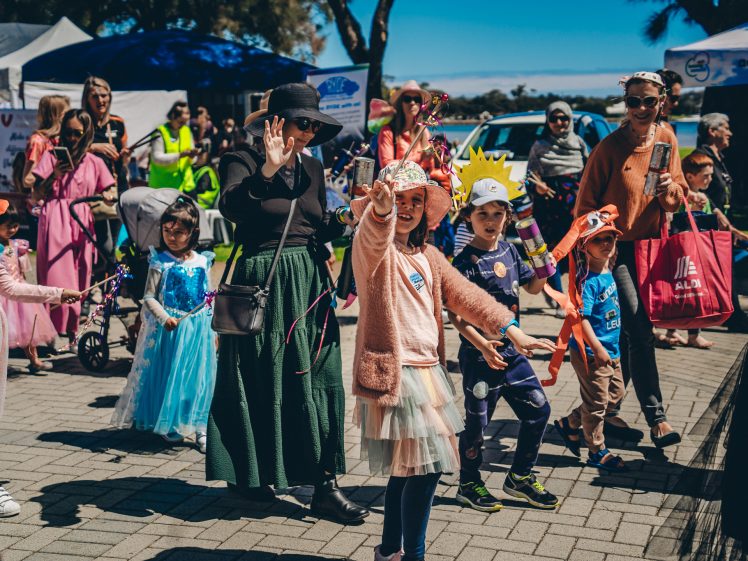 Image of A group of children and adults in colourful costumes enjoy an outdoor event, smiling and waving whilst standing on a paved area with tents and trees in the background.