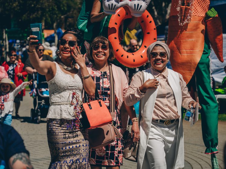Image of Three women smiling and posing for a selfie at an outdoor event, with colourful costumes and people in the background. The atmosphere is festive and sunny.