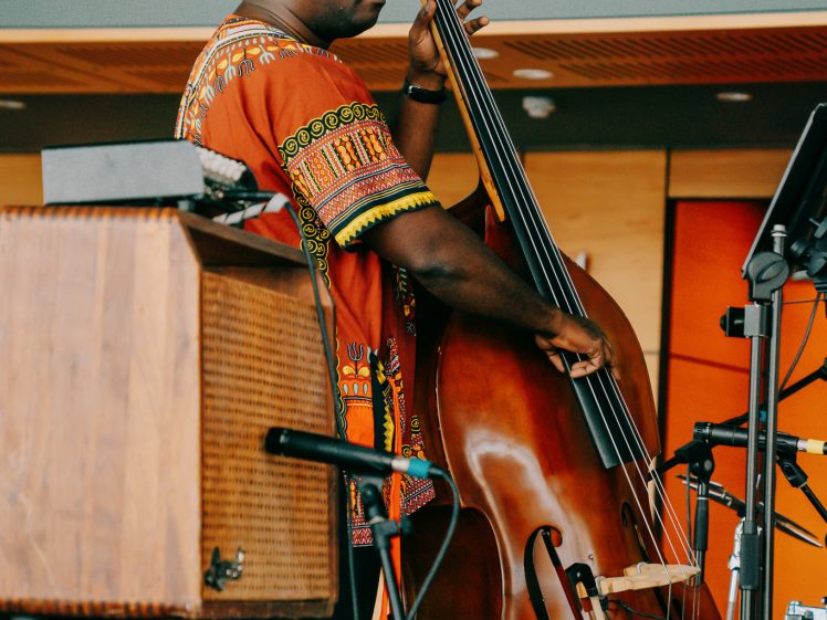 Image of A musician wearing a colourful patterned shirt plays a double bass on stage, surrounded by music equipment and microphones.