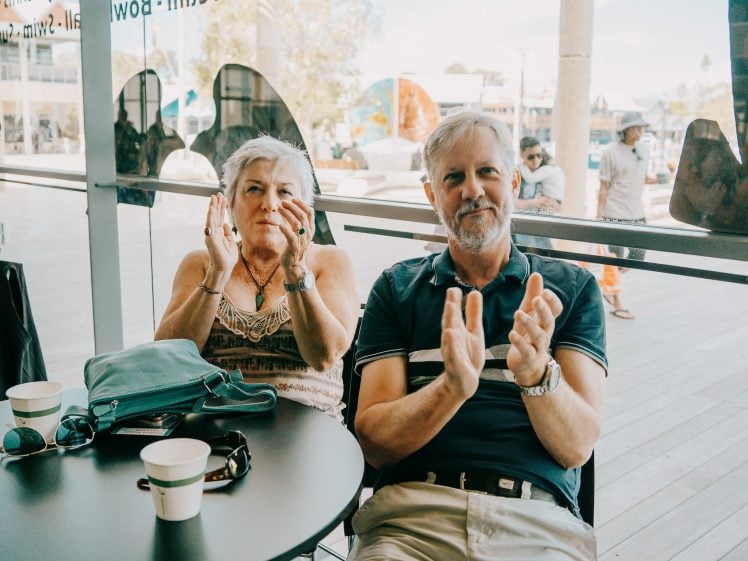 Image of An older woman and man sit at a café table with drinks, clapping and smiling. The background shows people walking outside through large glass windows.