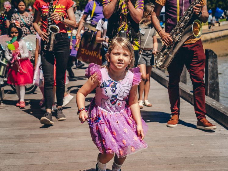 Image of A young girl in a pink dress jumps joyfully on a wooden path whilst a colourful street band plays instruments behind her; people are walking and enjoying the lively outdoor scene.