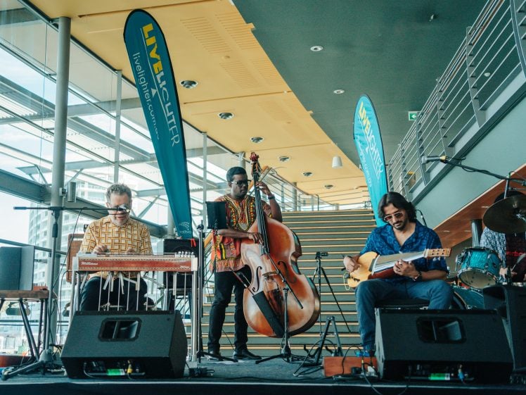 Image of Three musicians perform indoors: one plays a pedal steel guitar, another stands playing an upright bass, and the third plays an electric guitar. Banners and large windows are in the background.
