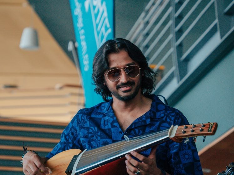 Image of A person wearing sunglasses and a blue patterned shirt plays a stringed instrument while sitting indoors near stairs, with a blue UNKASITA banner in the background.
