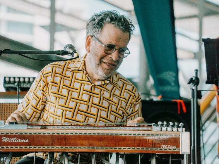 Image of An older man with glasses and a patterned shirt smiles while playing a Williams steel guitar at an indoor music event. A microphone and other musical equipment are visible in the background.