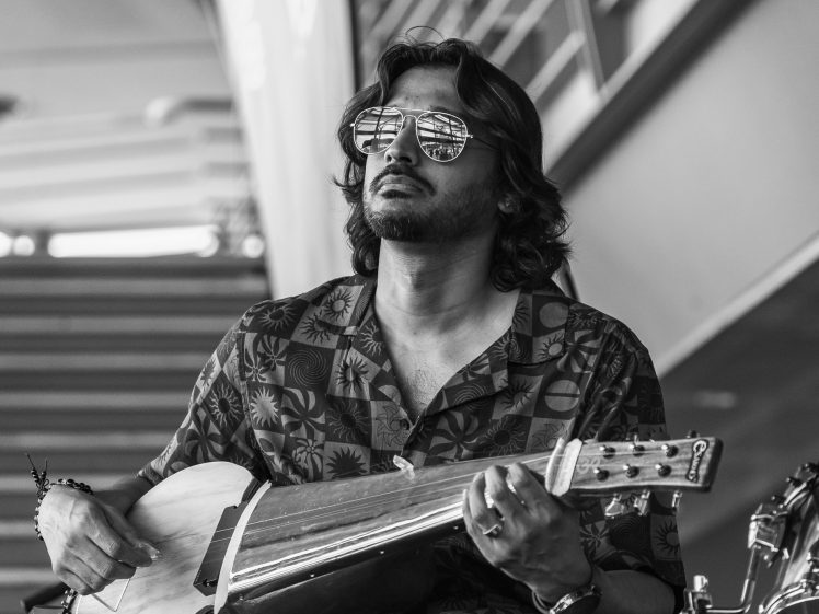 Image of A man with shoulder-length hair and sunglasses plays a stringed instrument. He wears a patterned shirt and a bracelet, and appears focused. The image is black and white with a blurred background.