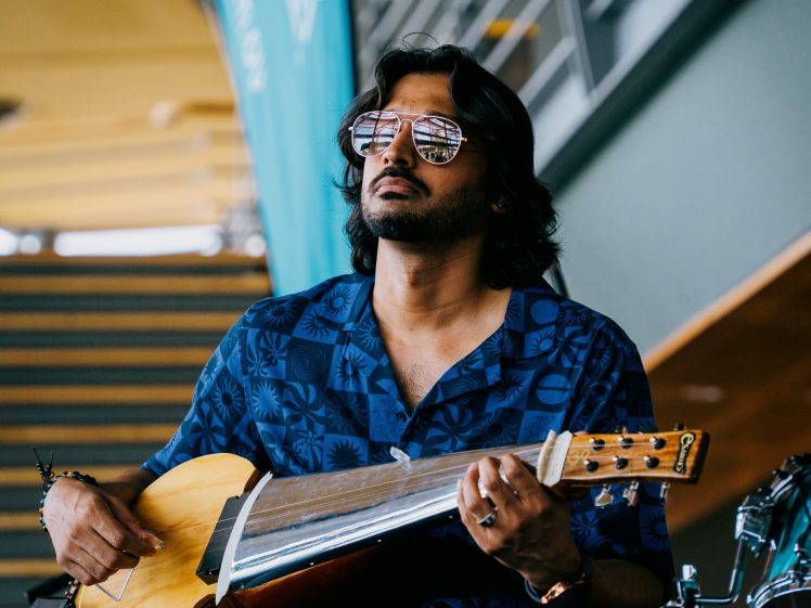 Image of A man wearing sunglasses and a blue patterned shirt plays a slide guitar. He appears focused, with long hair and facial hair, and is seated indoors near a staircase and green banner.