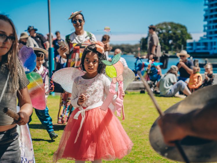 Image of A young girl in a pink skirt and fairy wings stands outdoors among other children and adults at a lively event, with colourful costumes, sunlight, and buildings in the background.