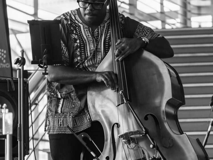 Image of A man wearing glasses and a patterned shirt plays a double bass in front of a staircase. The image is black and white, and he appears focused on his music, which is on a stand nearby.