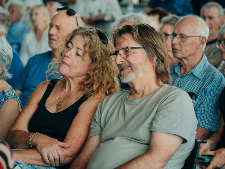 Image of A group of older adults sit closely together, listening attentively. In the foreground, a woman with curly hair leans on a man with glasses and a beard. Both appear relaxed and content in a crowded indoor setting.