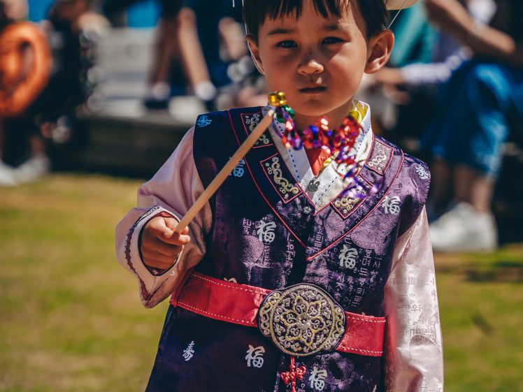 Image of A young boy wearing traditional Asian clothing and a white hat holds a colourful beaded wand, standing outdoors on grass with people sitting in the background.
