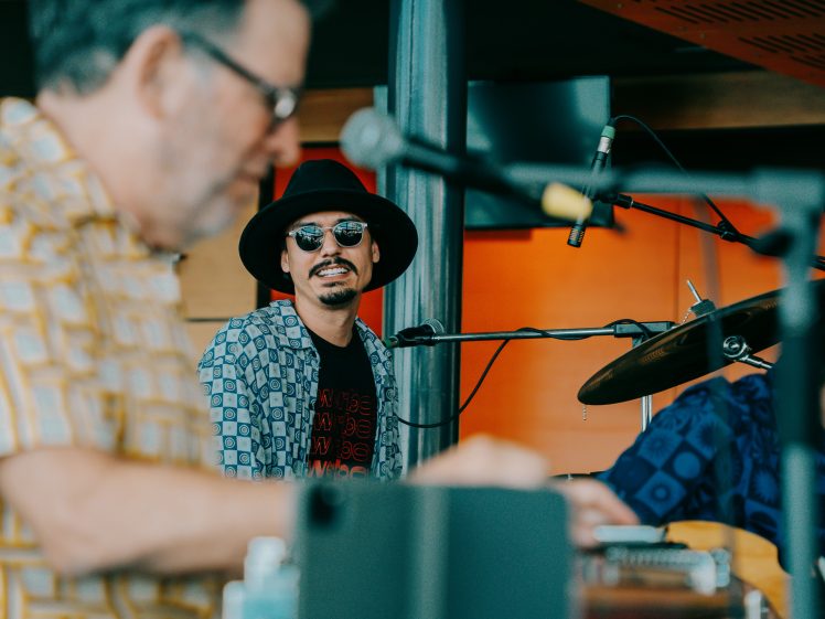 Image of A musician wearing sunglasses and a black hat sits at a drum kit, looking towards the camera, while another person in the foreground plays a keyboard. The background is warm-toned and out of focus.