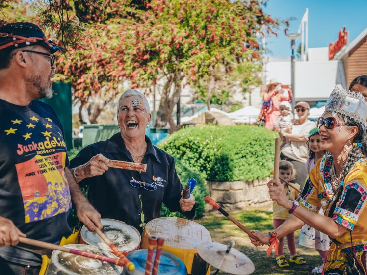 Image of A group of people joyfully play drums and percussion instruments outdoors, wearing colourful clothes and smiling, whilst others watch in the background at what appears to be a festive event.