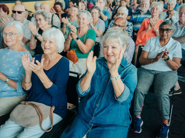 Image of A group of elderly people sit in rows of chairs, smiling and clapping. The atmosphere is cheerful, and most attendees wear casual clothes. The background shows several more people also applauding enthusiastically.