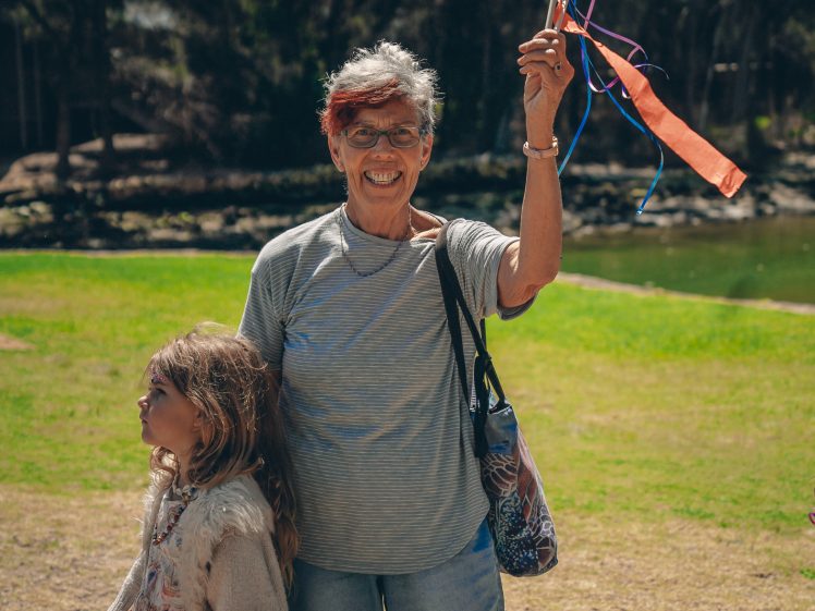 Image of An older woman smiles and raises a colourful ribbon wand whilst standing outdoors on grass beside a young girl in a dress and cardigan. Trees and water are visible in the background.