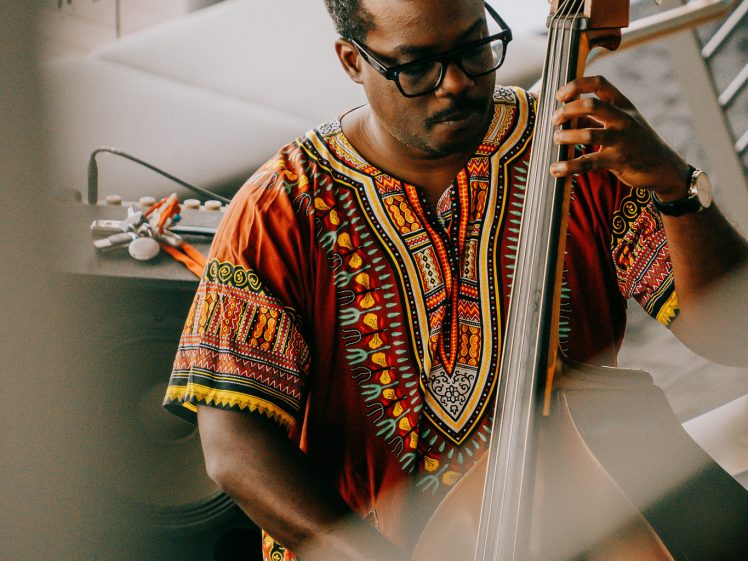Image of A man wearing a colourful traditional African shirt plays a double bass indoors, focusing intently on the instrument.