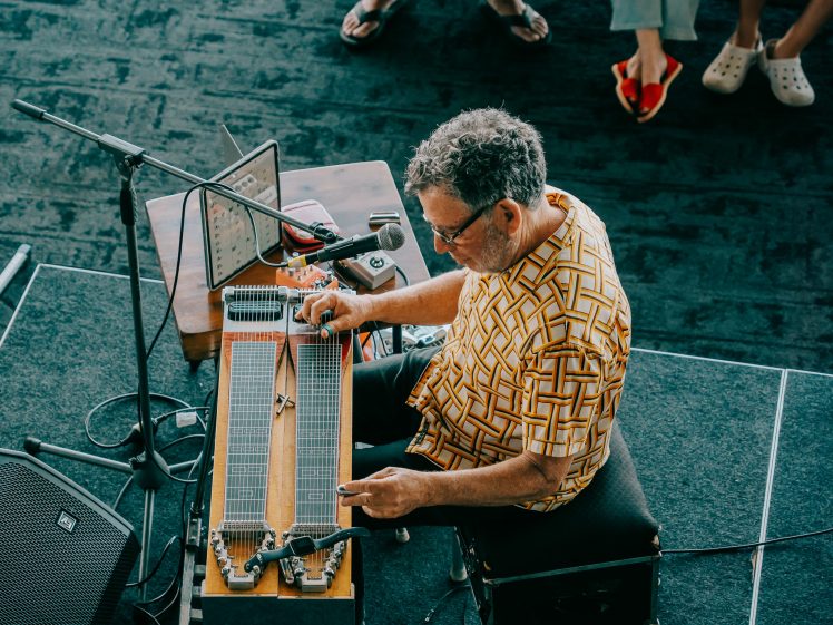 Image of A man in a patterned shirt plays a double-neck pedal steel guitar on stage. He is seated, focused, and surrounded by equipment. Audience members' legs and feet are visible in the background.