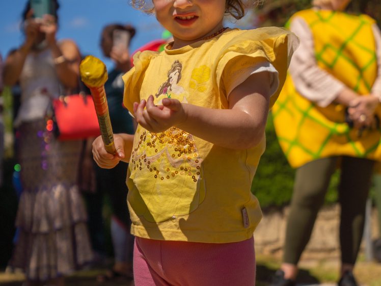 Image of A young child in a yellow shirt and pink trousers holds a toy wand and smiles outdoors, with adults in colourful costumes blurred in the background on a sunny day.