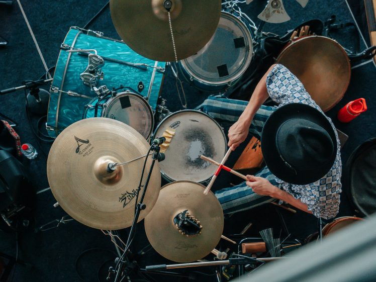 Image of Overhead view of a drummer wearing a black hat and patterned shirt, playing a drum kit with cymbals, drums, and percussion instruments. The drummer is holding red drumsticks.