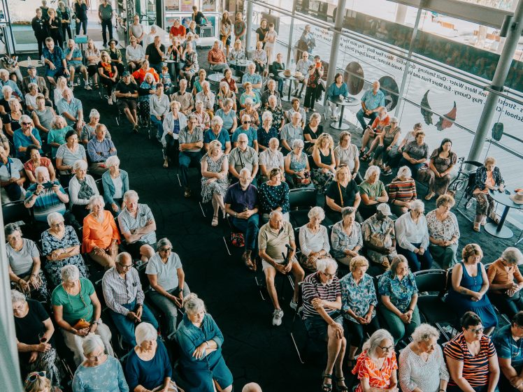 Image of A large group of people, mostly older adults, sit closely together in rows of chairs at an indoor event, viewed from above. Some people stand or sit at tables along the windows. The atmosphere appears social and engaged.