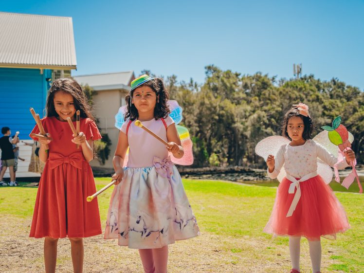 Image of Three young girls in colourful dresses and wings stand outdoors on a sunny day, holding wands and dressed as fairies. A house, trees, and a person in the background complete the cheerful scene.