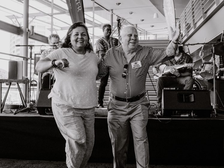 Image of Two smiling older adults dance together in front of a stage with a live band. Several musicians play instruments in the background. The scene appears lively and joyful in a spacious indoor venue.