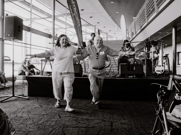 Image of Two smiling older adults dance hand in hand in front of a stage with live musicians, inside a bright, modern venue with large windows. Other people are seated, watching the performance. The scene feels joyful and lively.