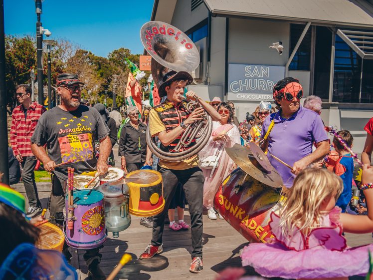 Image of A lively street band plays music outdoors, surrounded by people in colourful costumes and masks. Children and adults enjoy the festive atmosphere near a building labelled “San Churro Chocolateria.”.