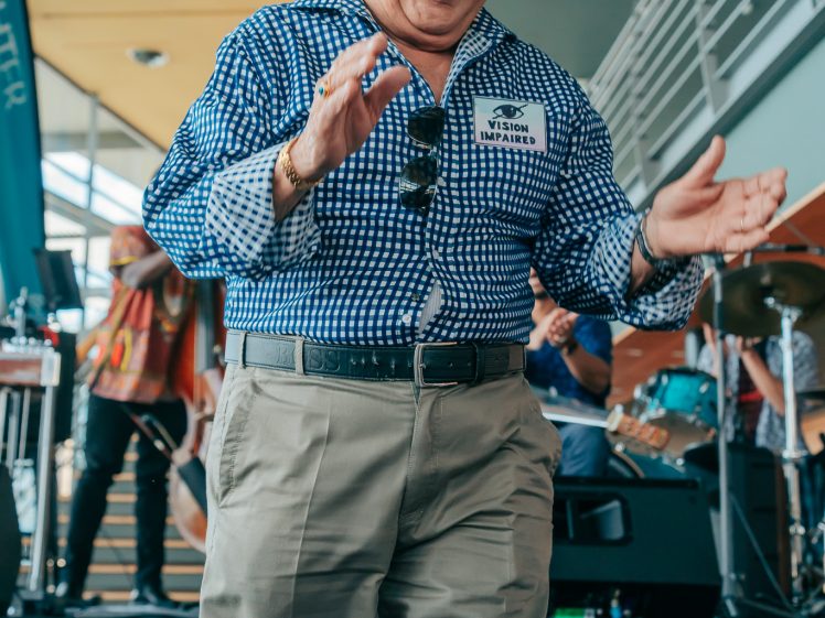 Image of An older man with short white hair, wearing a blue chequered shirt and khaki trousers, smiles while dancing with his eyes closed. He is indoors, near a stage with musical instruments in the background.