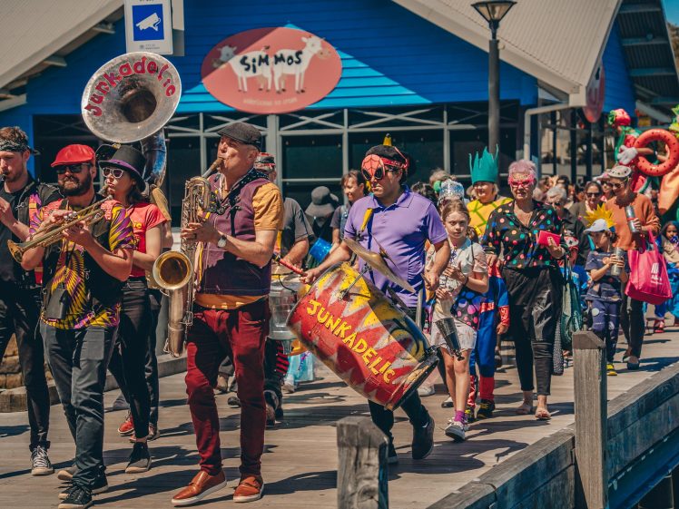 Image of A lively band in colourful outfits leads a cheerful parade with people of all ages in fancy dress, marching along a promenade with a blue building and a sign reading Simmos in the background.