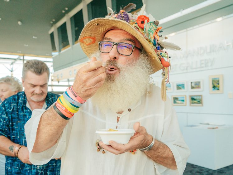 Image of A bearded man wearing a colourful, decorated hat and glasses eats from a small white bowl at an art gallery event. People are standing behind him and artwork hangs on the walls.