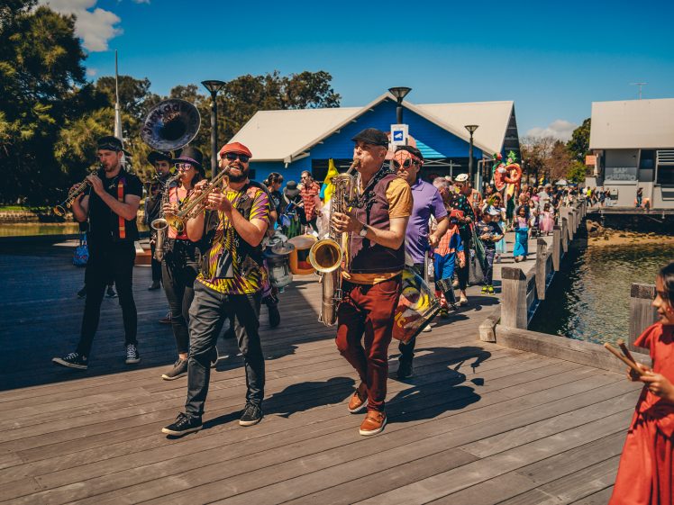 Image of A lively parade with musicians playing brass instruments walks along a wooden promenade by the water, followed by a festive crowd under a bright blue sky. Some people wear colourful clothes, and buildings are visible in the background.