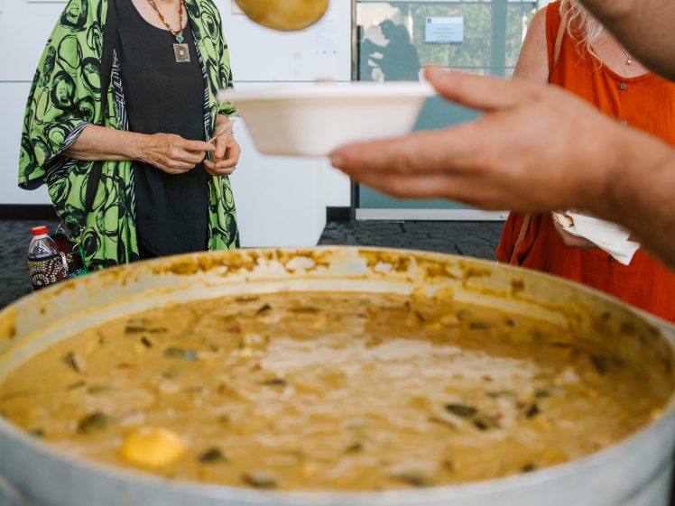 Image of A large pot of soup is being ladled into a bowl by a person, while a smiling woman in a green patterned jacket and black hat stands nearby. The scene appears to be at a community event or gathering.