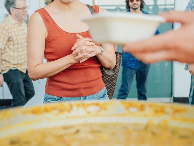 Image of A woman in an orange vest stands with her hands clasped, watching as someone ladles food into a bowl from a large pot. People and a sign are visible in the blurred background.