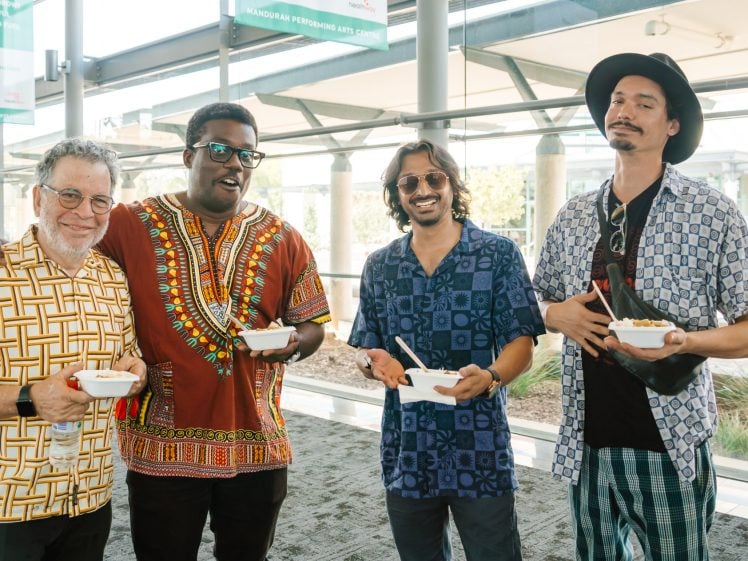 Image of Four men stand indoors, smiling at the camera whilst holding bowls of food. They wear colourful, patterned shirts and sunglasses. Sunlight streams through large windows in the background.