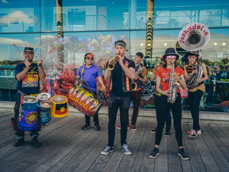 Image of A colourful street band with six musicians plays various instruments, including drums, saxophones, and a sousaphone, in front of a glass building with graffiti art. The performers wear bright, eclectic clothing and accessories.