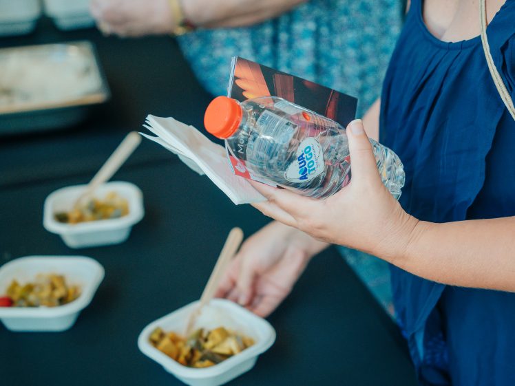 Image of A person in a blue top holds a bottle of water, serviettes, and a booklet whilst standing by a table with bowls of food and wooden cutlery.